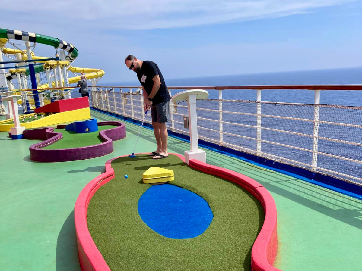 Man playing miniature golf on a cruise ship deck, aiming a putt on colorful artificial turf with the ocean in the background