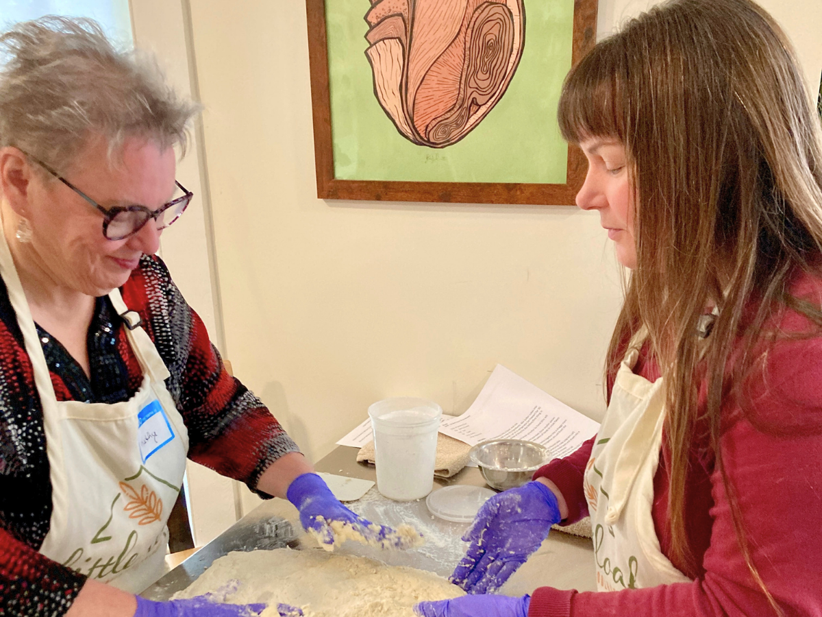 Two women in aprons kneading dough on a flour-dusted counter, wearing purple gloves and smiling at each other during a baking workshop.