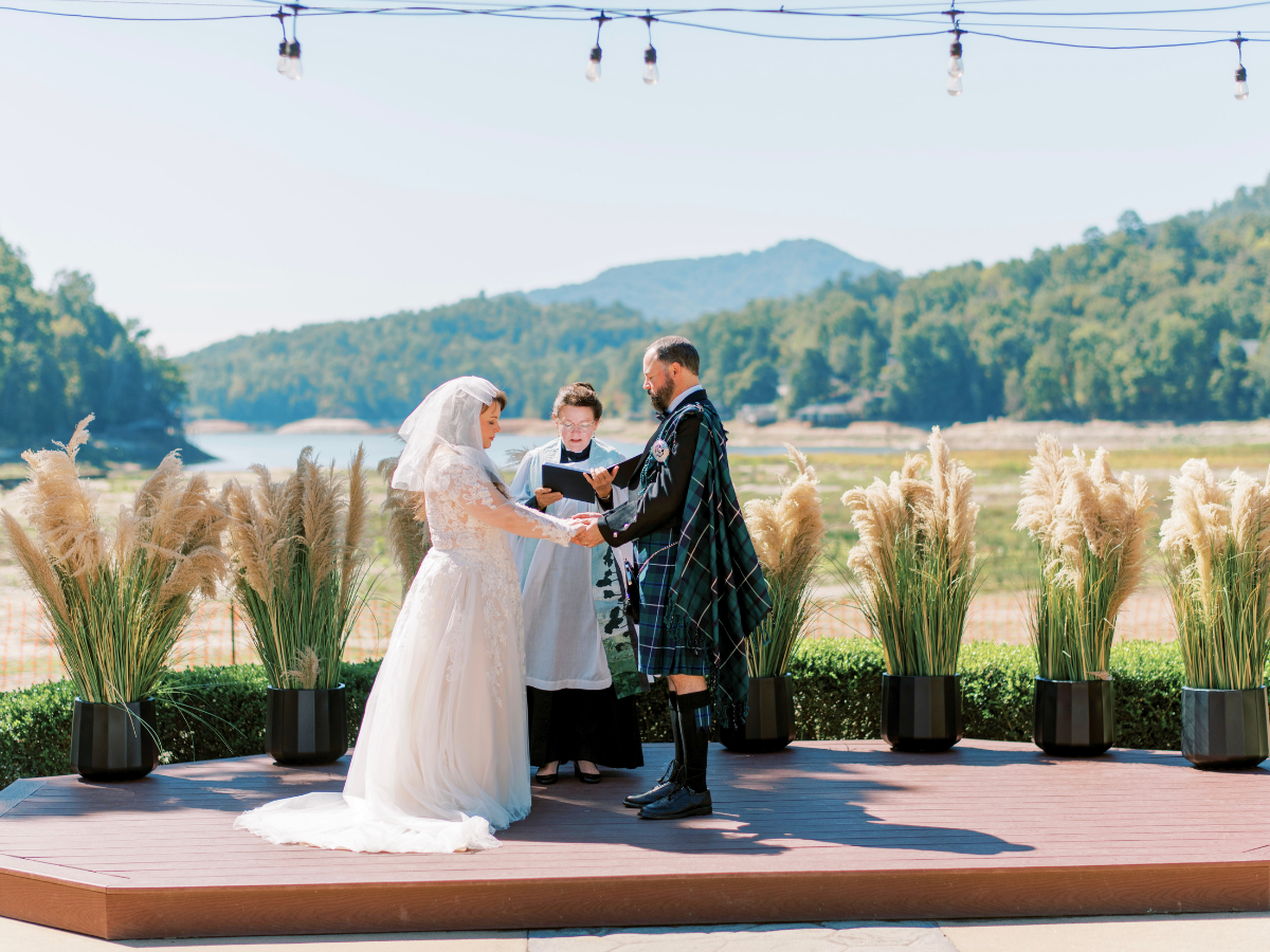 Bride in a lace gown and groom in a plaid kilt exchange vows on a wooden platform by a lake, with an officiant and tall pampas grasses.