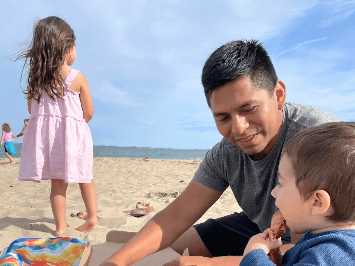 Father with two kids on a sandy beach; girl in pink dress stands near the water while boy eats a snack nearby.