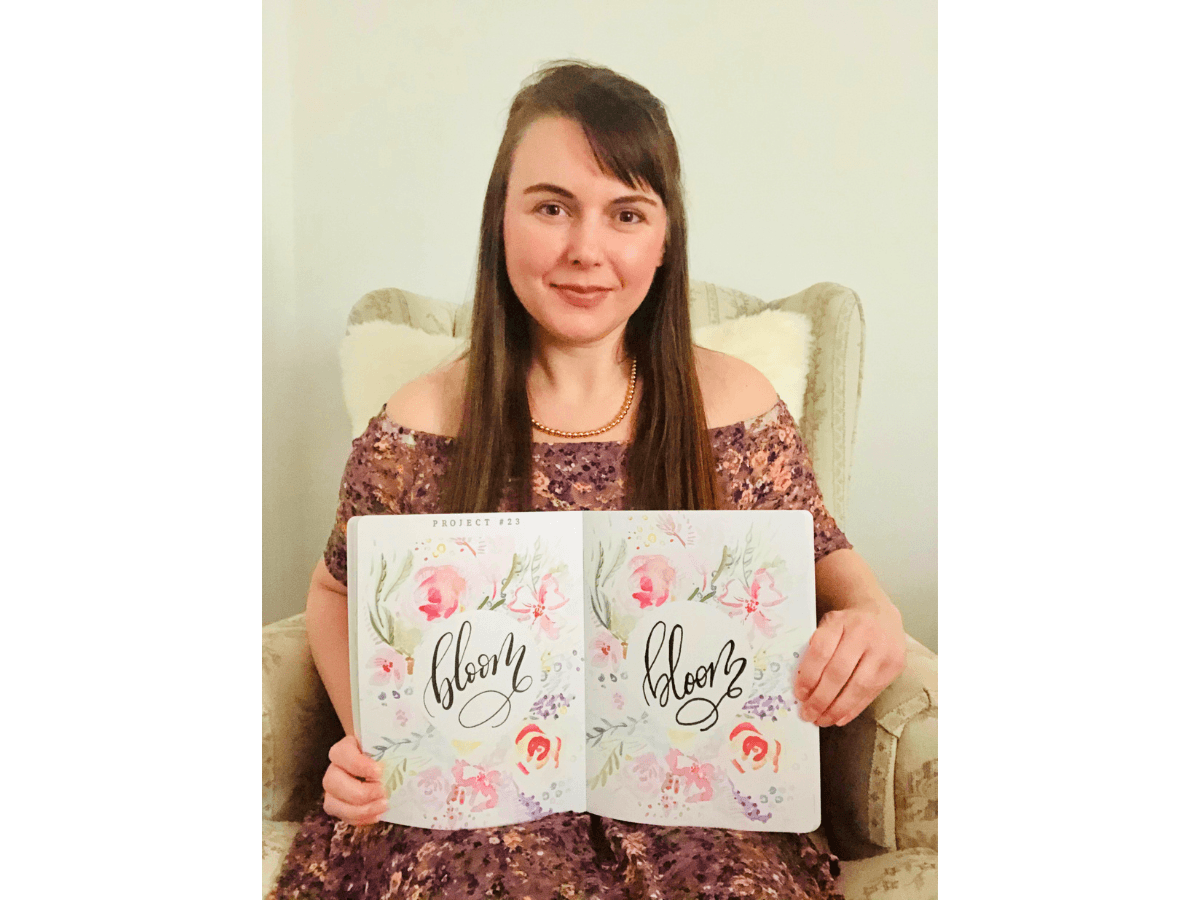 Woman in a floral off-shoulder dress sits in a cream chair, holding two open floral journals that read 'Bloom'.