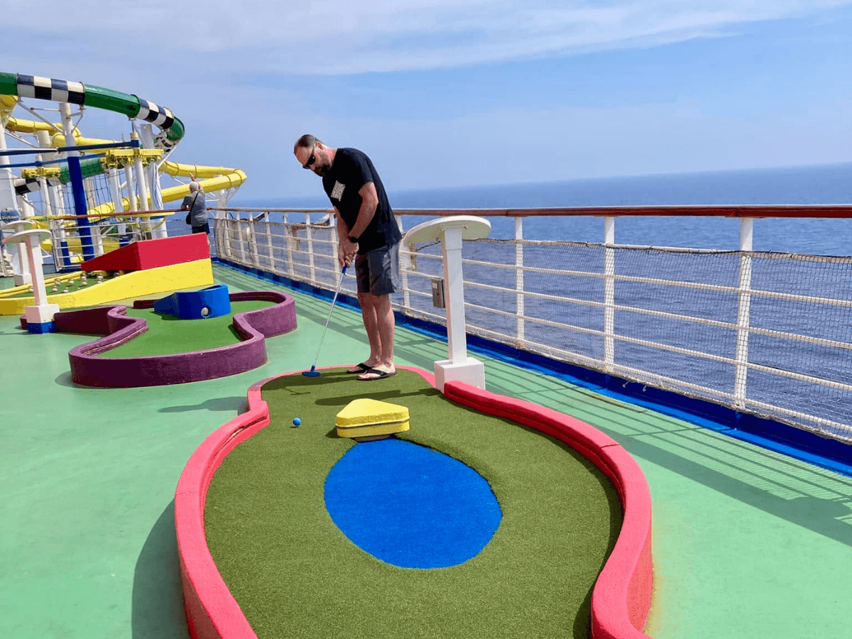 Person wearing a black shirt and shorts playing mini-golf on a ship deck, with colorful obstacles and the ocean in the background.