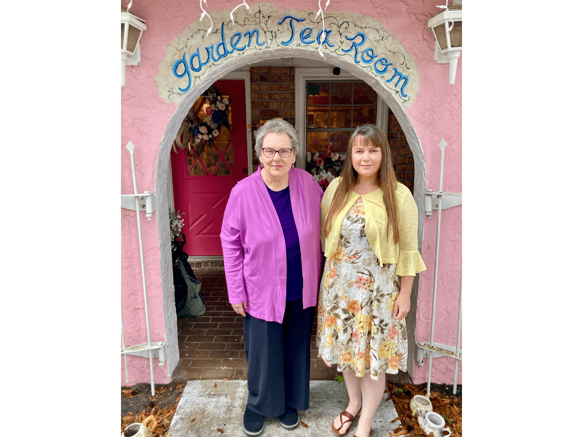 Two women pose in front of a pink building with a curved sign reading 'Garden Tea Room' above the entrance.