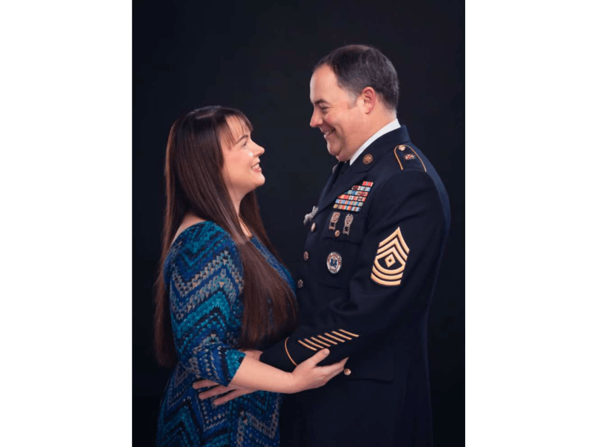 Smiling woman in a blue patterned dress stands close to a smiling man in a decorated military uniform.