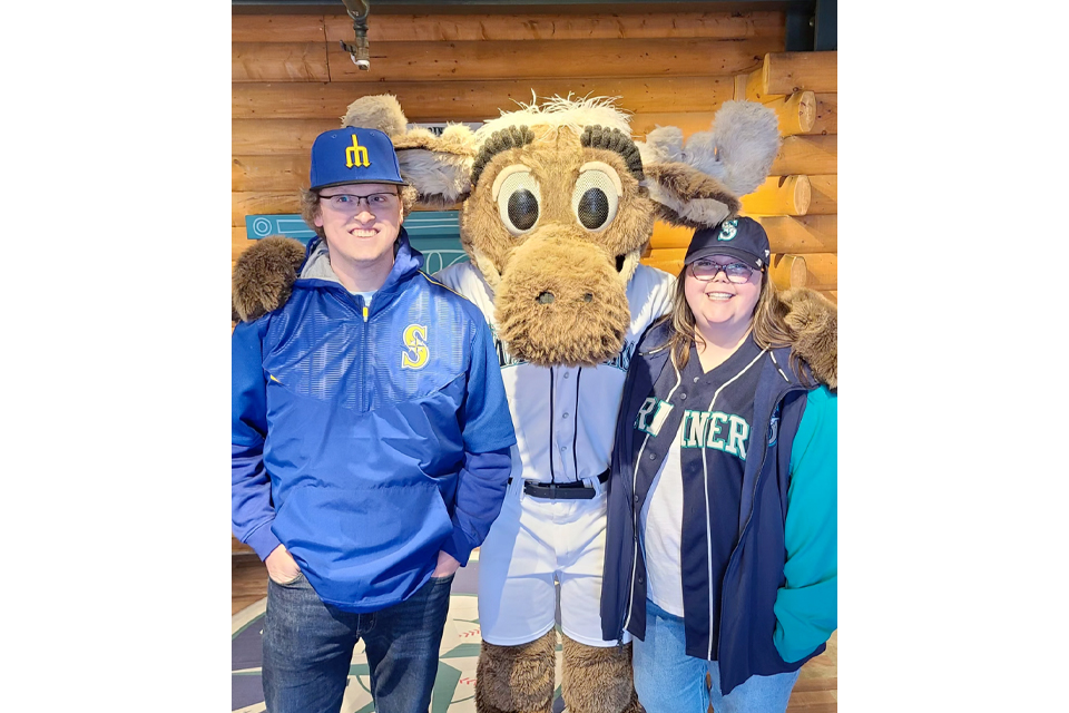 Two fans pose with a large moose mascot between them inside a log-cabin style venue, all smiling for the photo.