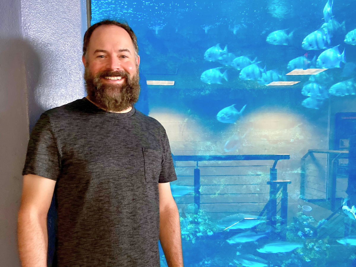 Smiling man with a beard in a dark gray T-shirt stands beside a large blue aquarium filled with swimming fish.