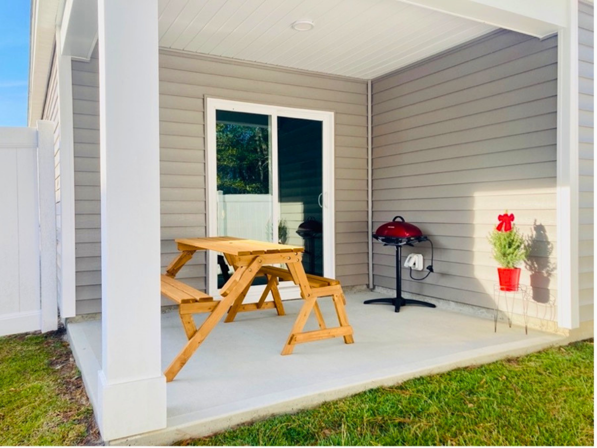 A small covered patio with a wooden picnic table and a red kettle grill, beige siding, a sliding glass door, and a potted plant with a red bow on the right.
