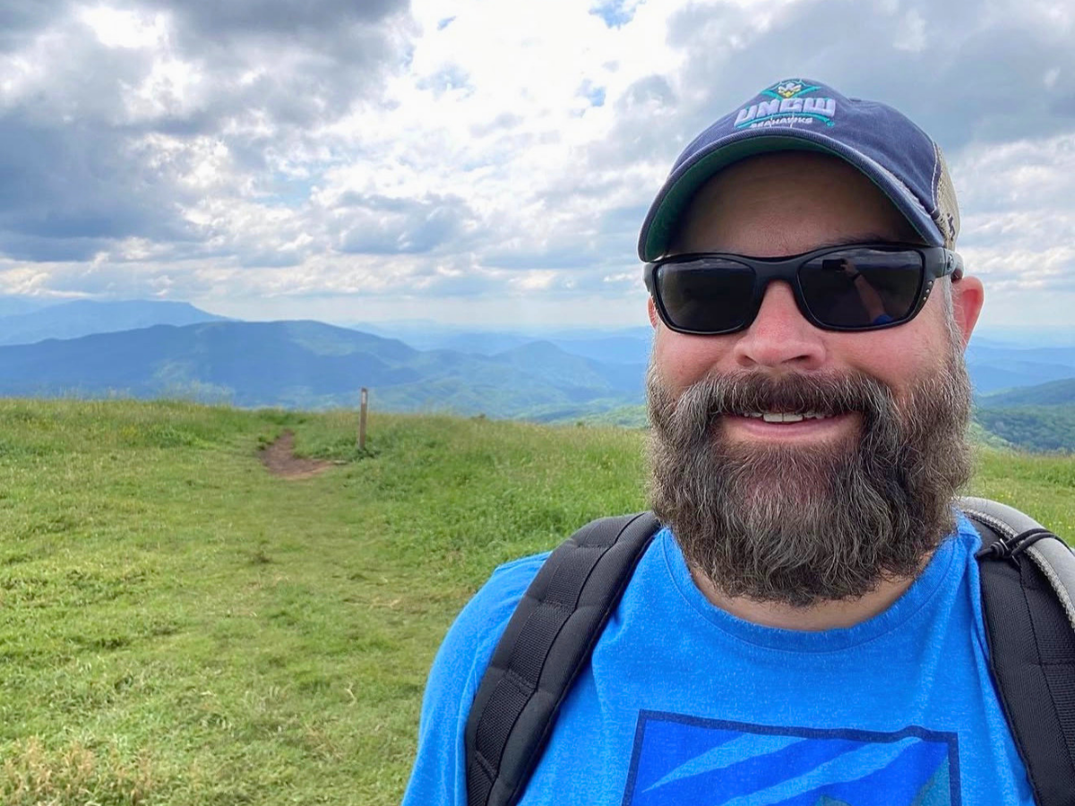 Man with a beard wearing black sunglasses and a blue t-shirt, taking a selfie on a grassy hill with mountains in the distance and a backpack on his shoulders