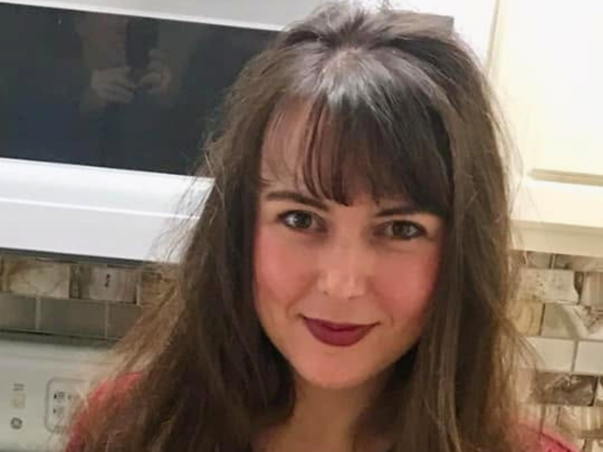 Portrait of a smiling woman with long dark hair and bangs in a kitchen setting, wearing a red top.