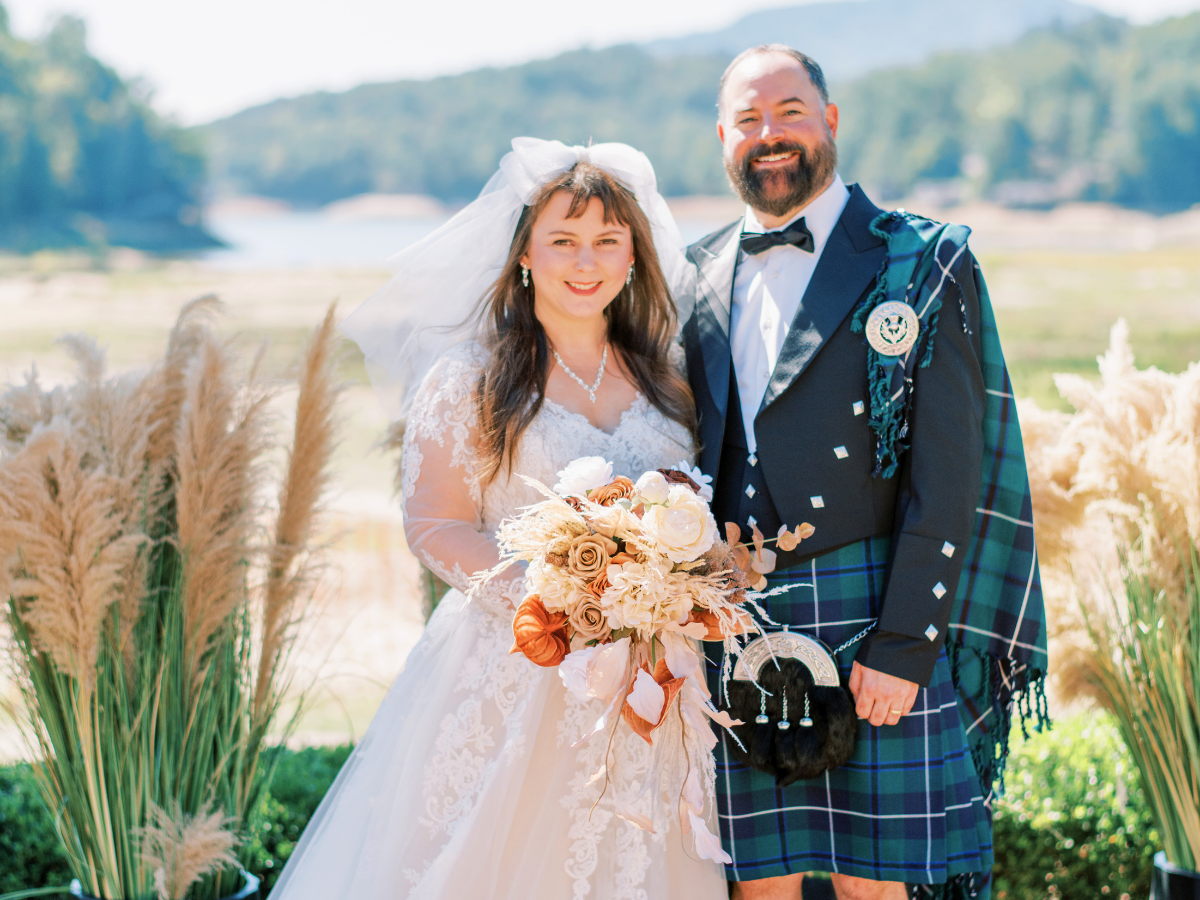 Bride in a lace wedding gown and veil holds a cream bouquet beside a man in a blue plaid kilt outdoors, both smiling.