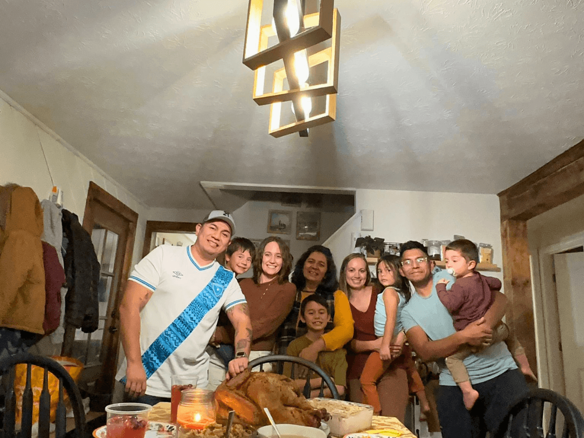 Family and friends gathered around a dining table with a roasted turkey, posing for a group photo in a cozy dining room.