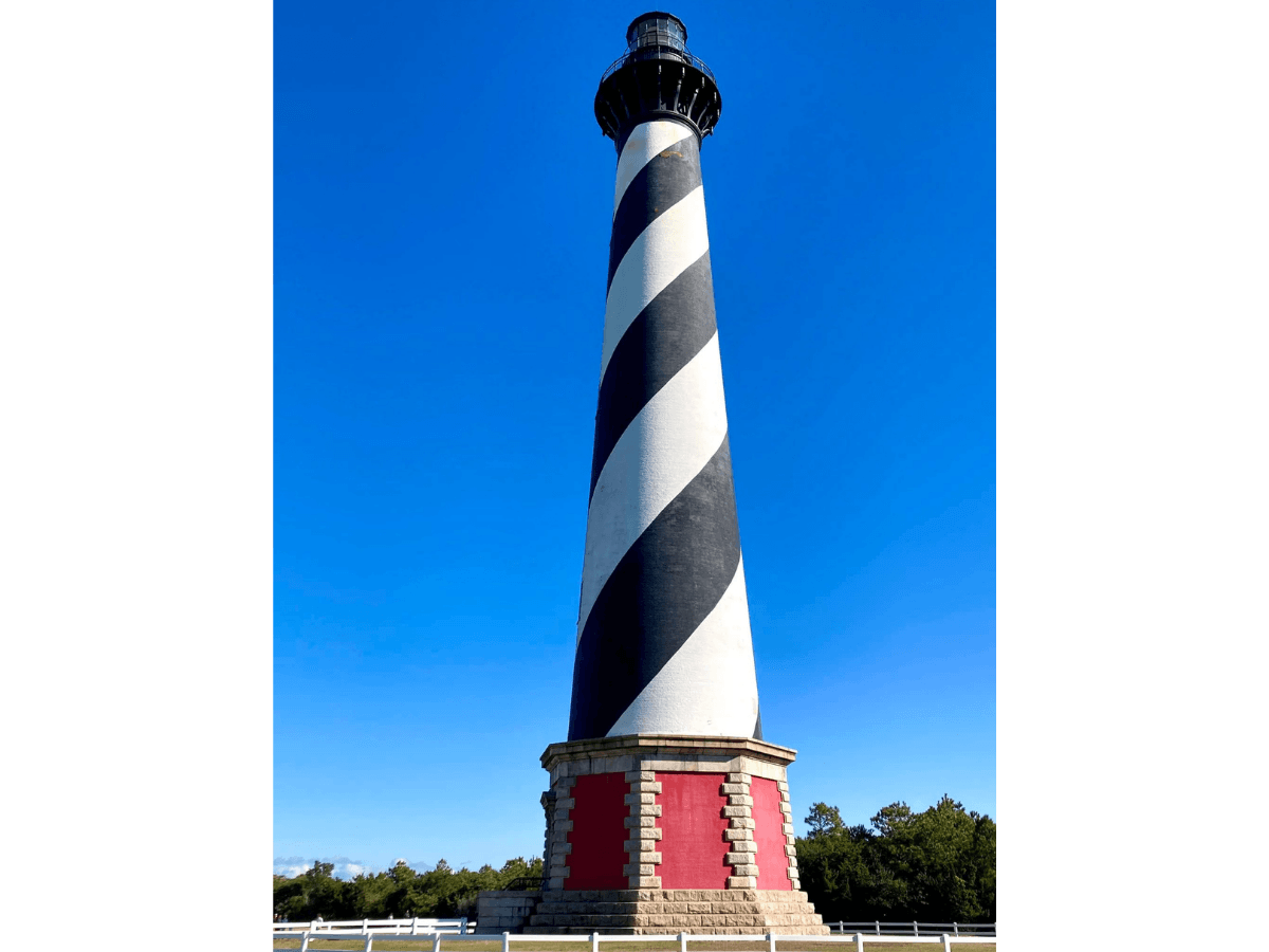 Tall lighthouse with bold diagonal black-and-white stripes and a red base against a clear blue sky.