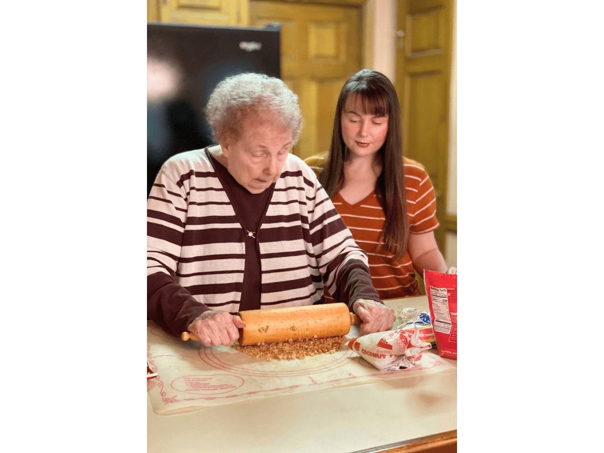 Older woman rolling dough with a rolling pin while sprinkled nuts spill on the table; a younger woman watches nearby in a kitchen.