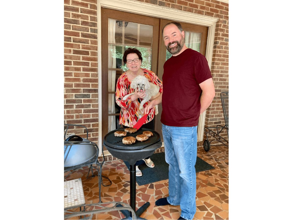 Couple grilling burgers on a patio; woman holds a small white dog while man smiles nearby.