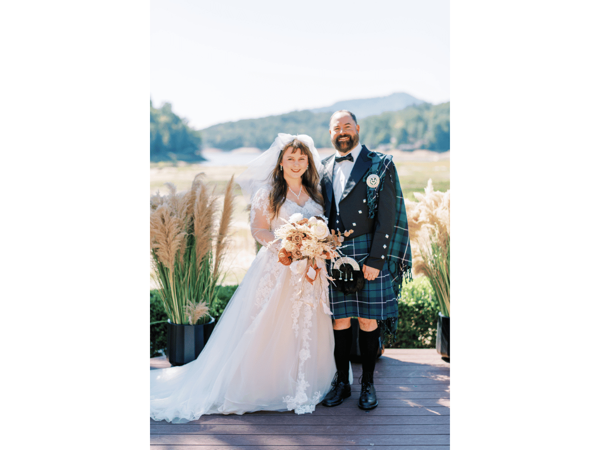 Bride and groom pose outdoors on a wooden deck; bride holds a cream bouquet, wearing a lace gown and veil, while the groom wears a tartan kilt and jacket.