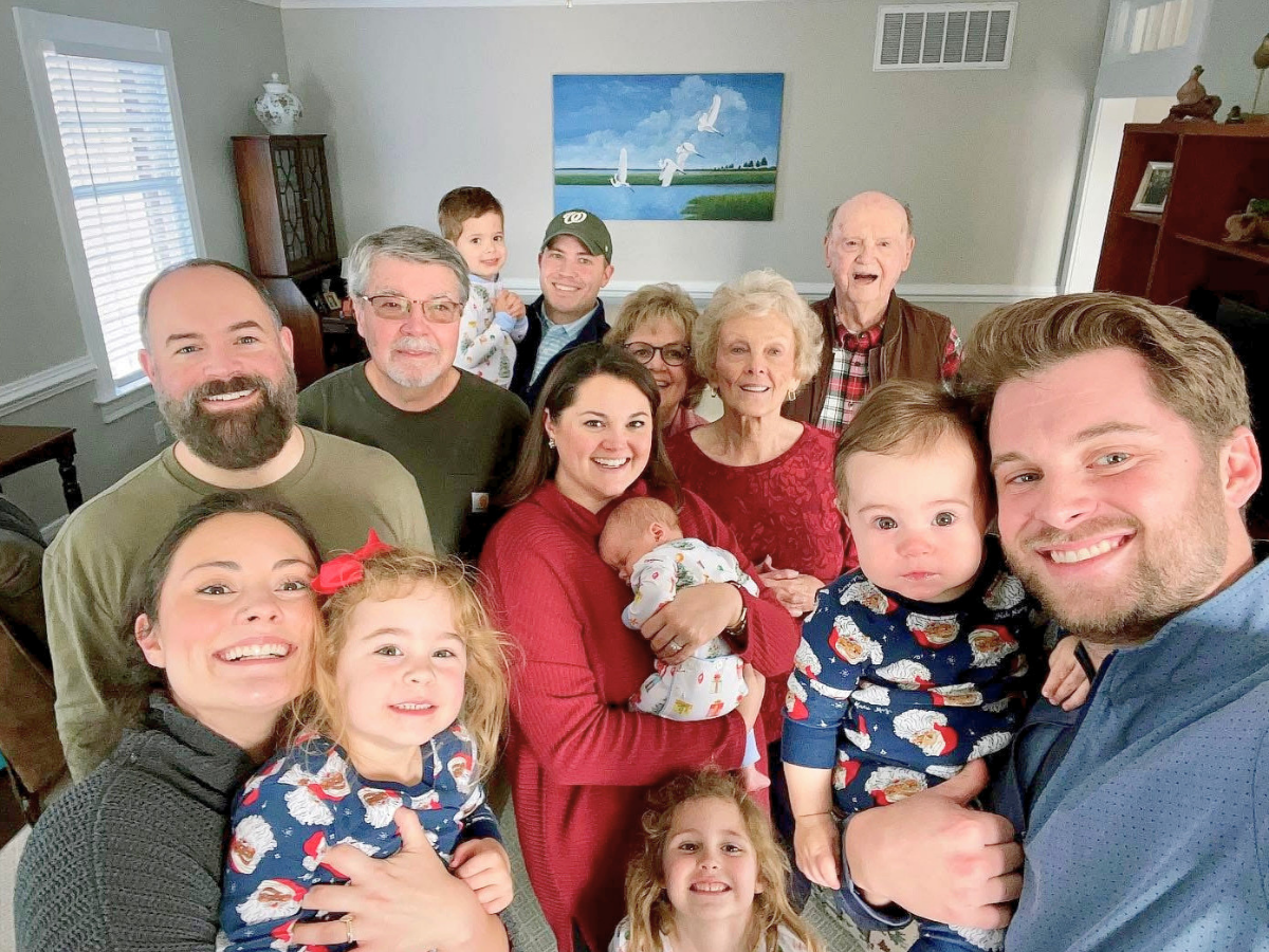 Multigenerational family portrait posing for a group selfie in a bright living room, smiling adults, seniors, and kids, with a baby in arms and children in festive pajamas