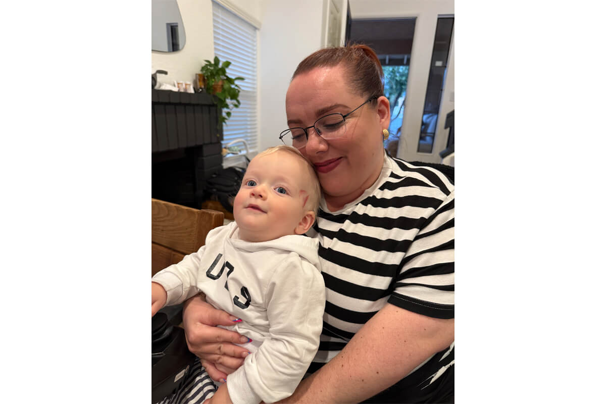Mother with glasses holds a smiling baby in a white hoodie at home, in a cozy room with a plant in the background.