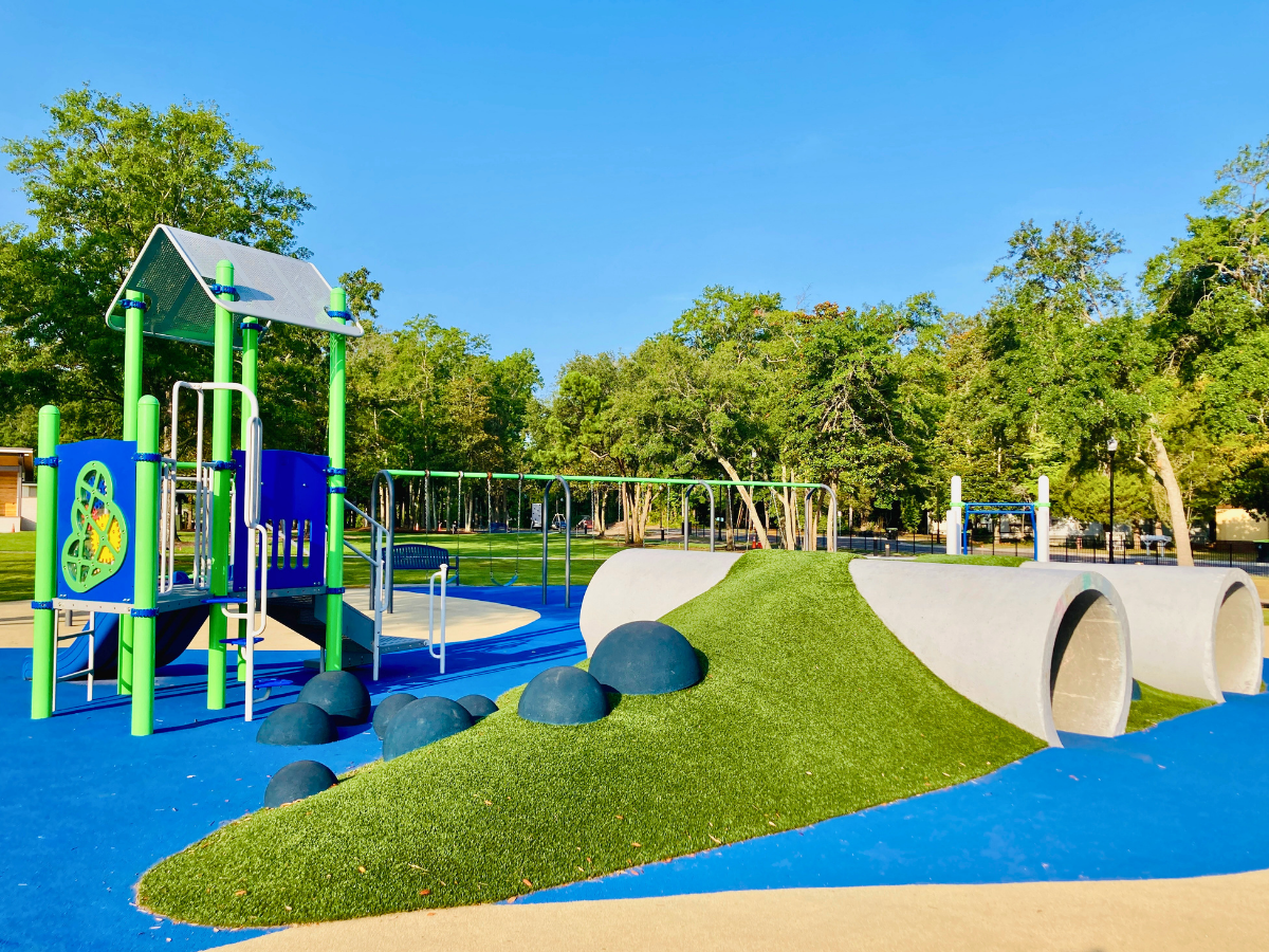 Colorful playground with a green and blue climbing structure, blue rubber ground, artificial grass hill, and curved concrete tube tunnels.