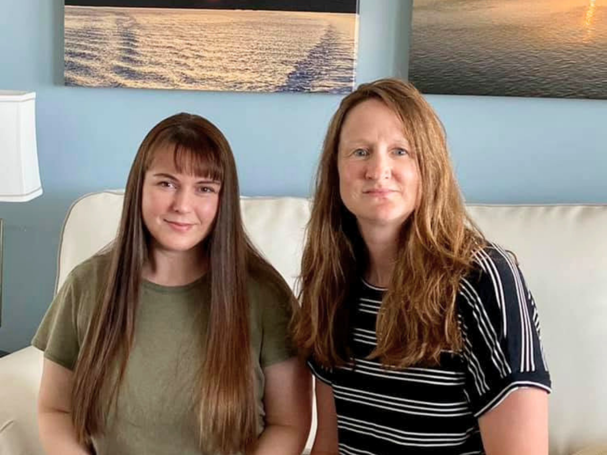 Two women sit together on a pale sofa in a blue-walled living room, smiling at the camera.