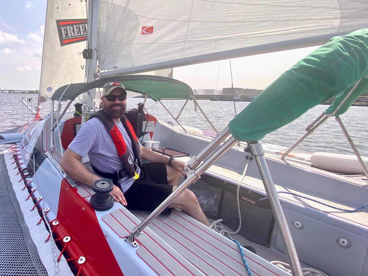 Man sailing a small racing sailboat, wearing a gray shirt, red life vest, cap, and sunglasses on the cockpit bench by the water.