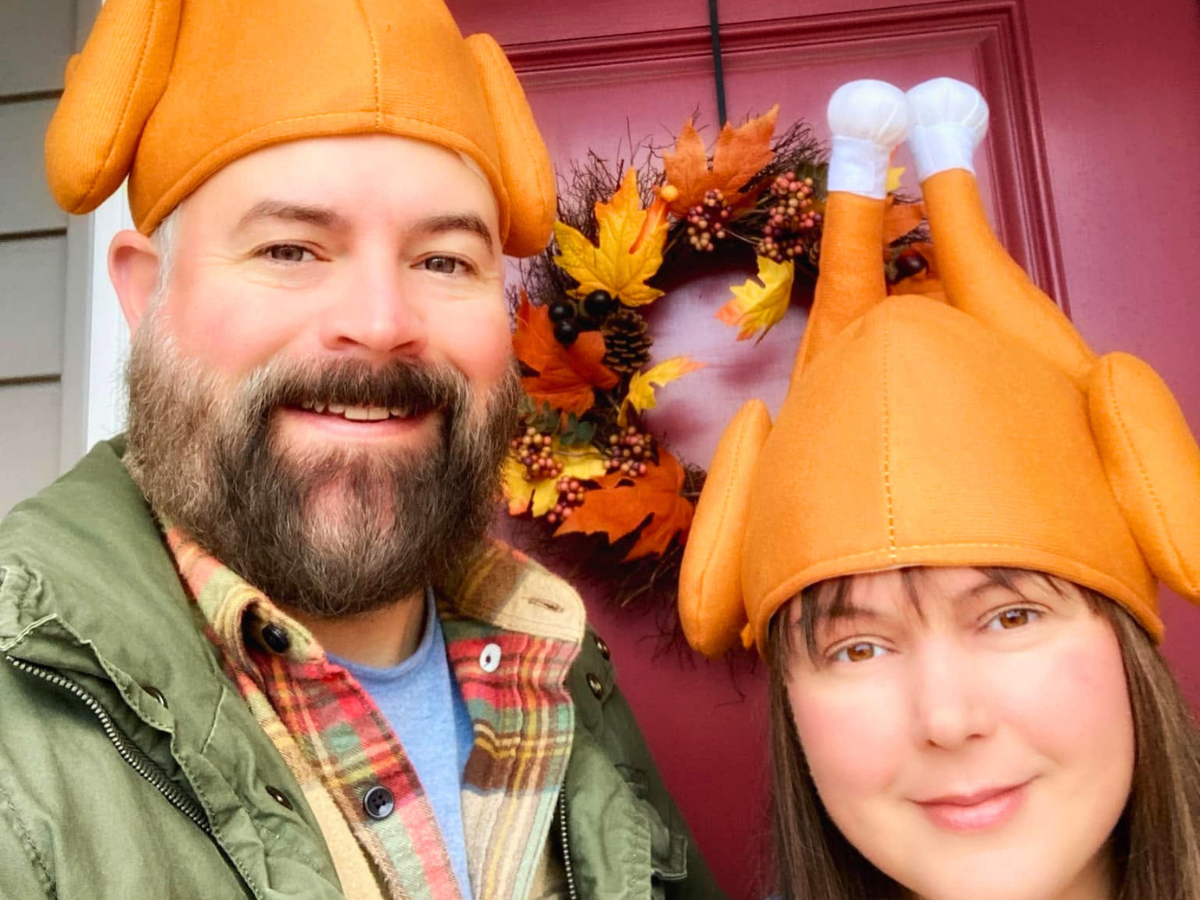 Two people wearing orange turkey hats smile for a selfie at a fall front door with a wreath behind them edge of a porch.