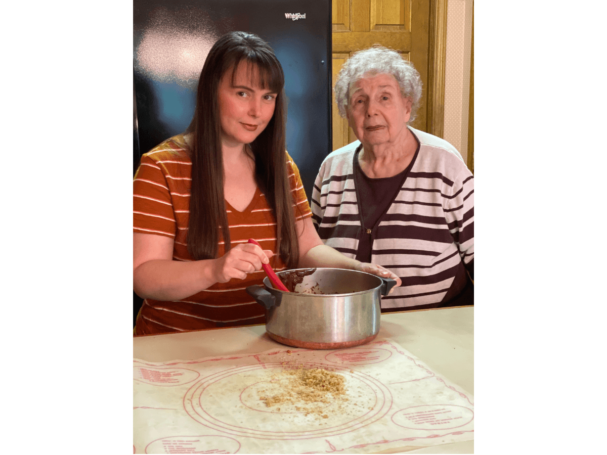 Young woman and older woman cooking together at a kitchen table, stirring a pot with a red spatula.