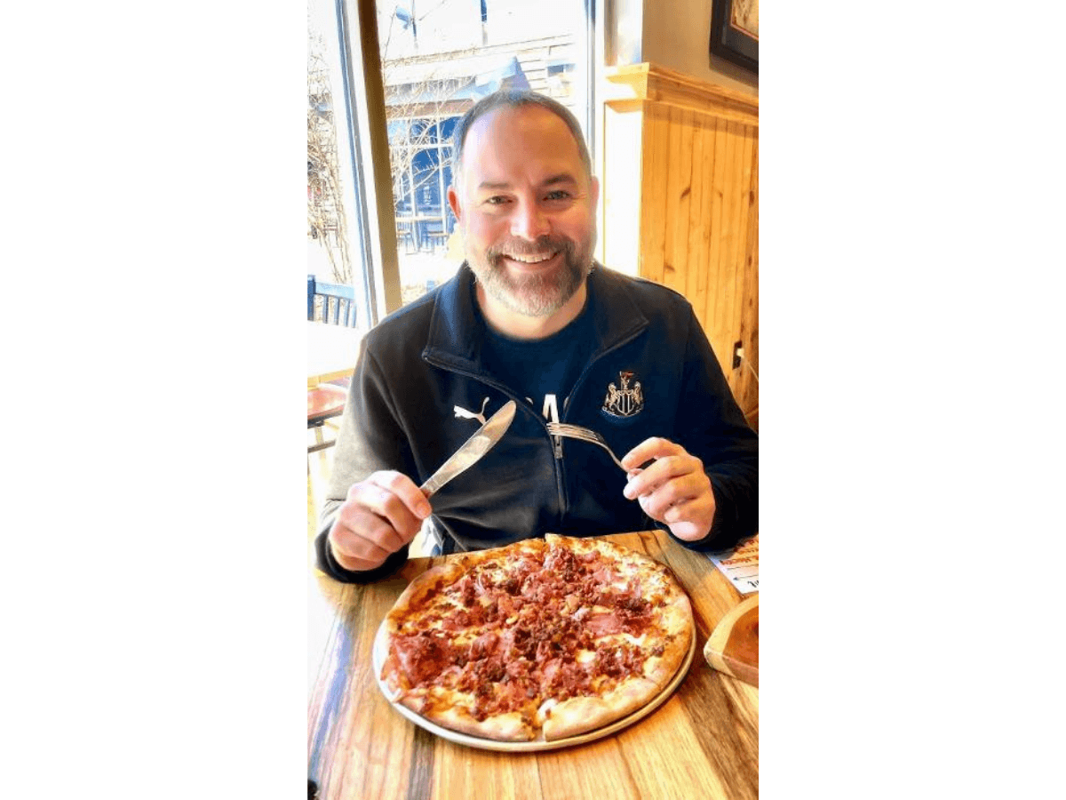 Smiling man in a dark jacket sits at a wooden table in a restaurant, about to eat a pepperoni pizza with a knife and fork.