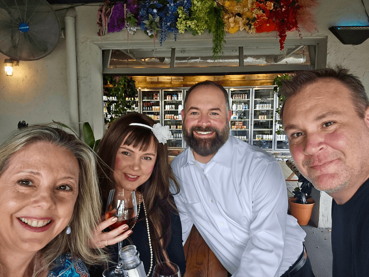 Four friends pose for a selfie at a bar, smiling; one holds a wine glass, with colorful flowers above and beverage shelves behind them.