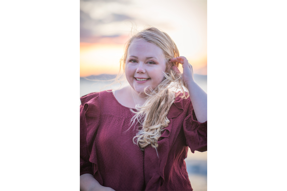Smiling woman with long blonde hair in a burgundy blouse at sunset on a beach.