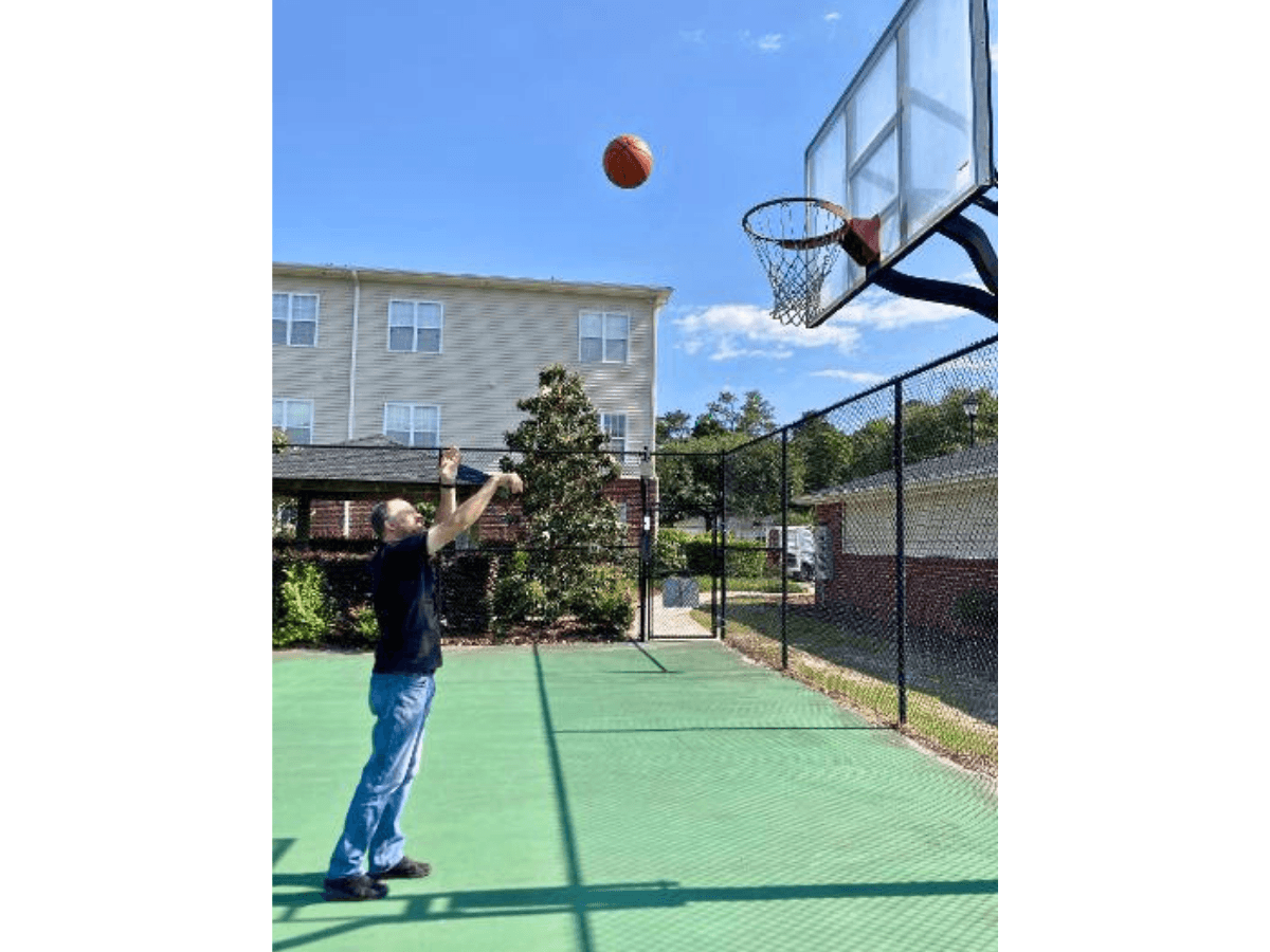 Man shooting a basketball toward a hoop on an outdoor court with a chain-link fence and apartment buildings in the background.