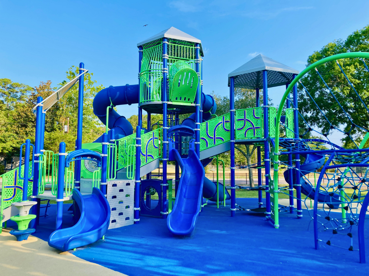 Colorful blue and green playground with enclosed tube slides, ladders, and climbing nets in a sunny park.