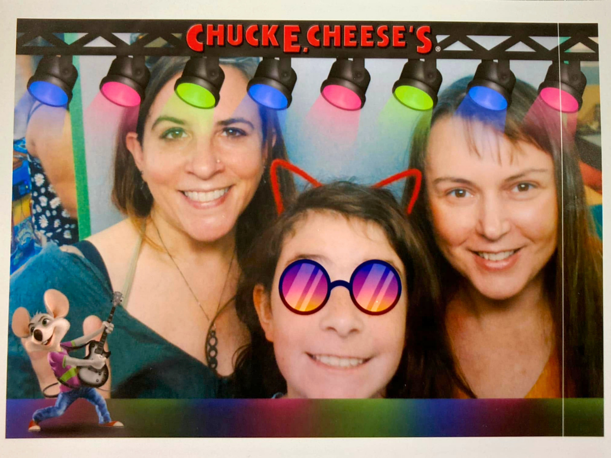 Three smiling friends take a selfie at Chuck E. Cheese's under colorful overhead lights, with a cartoon mouse and playful filters.