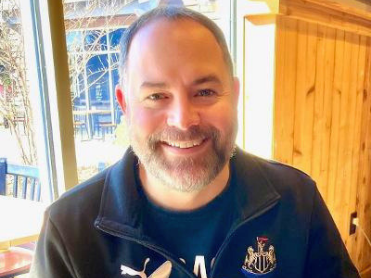 Smiling man with short hair and beard inside a cafe, wearing a dark jacket with crest on chest.