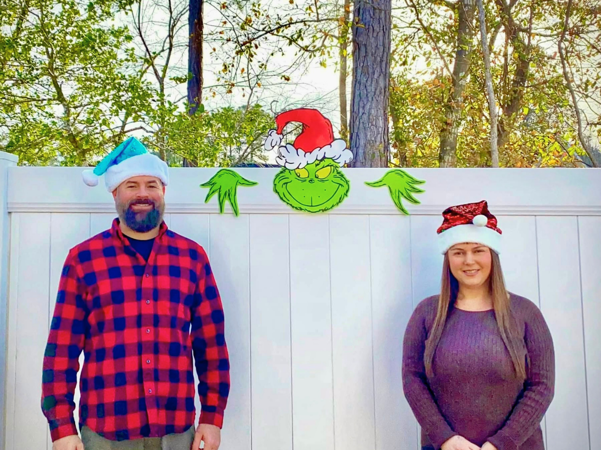 Two adults wearing festive Santa hats stand in front of a white fence, with a Grinch face and green decorations visible above them in a yard scene.