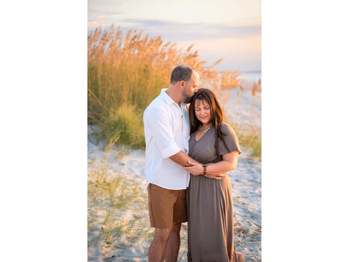 Couple embracing on a sandy beach at sunset, the man kissing the woman's head as they hold hands.