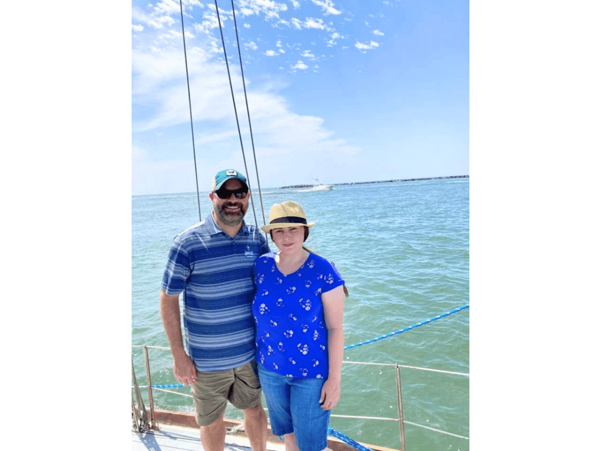 Two adults standing close together on a sailboat deck, smiling, with the blue ocean and clear sky in the background. The man wears a striped polo and a green cap; the woman wears a blue floral shirt and a straw hat.