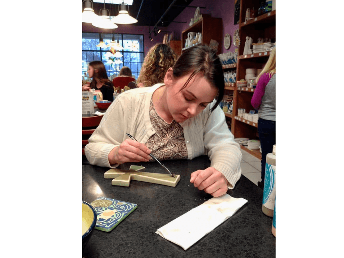 Woman focused on a craft project at a store counter, carving a light-colored cross-shaped piece with a tool.