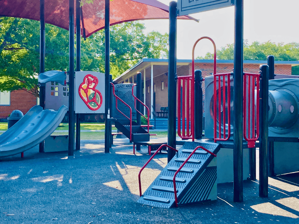 Colorful playground structure with blue slides, red railings, stairs, and a tubular tunnel in a sunny park setting.