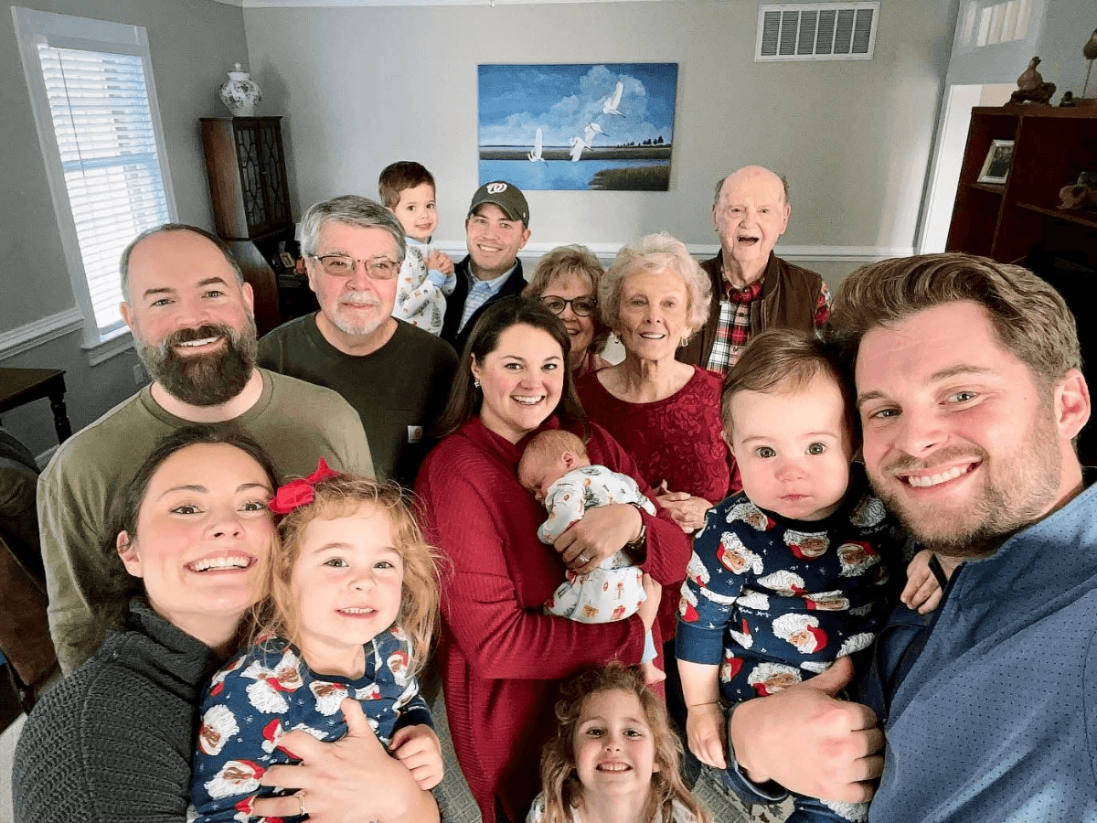 Large multi-generational family poses for a group portrait in a living room, smiling together; several adults, kids, and two babies present.
