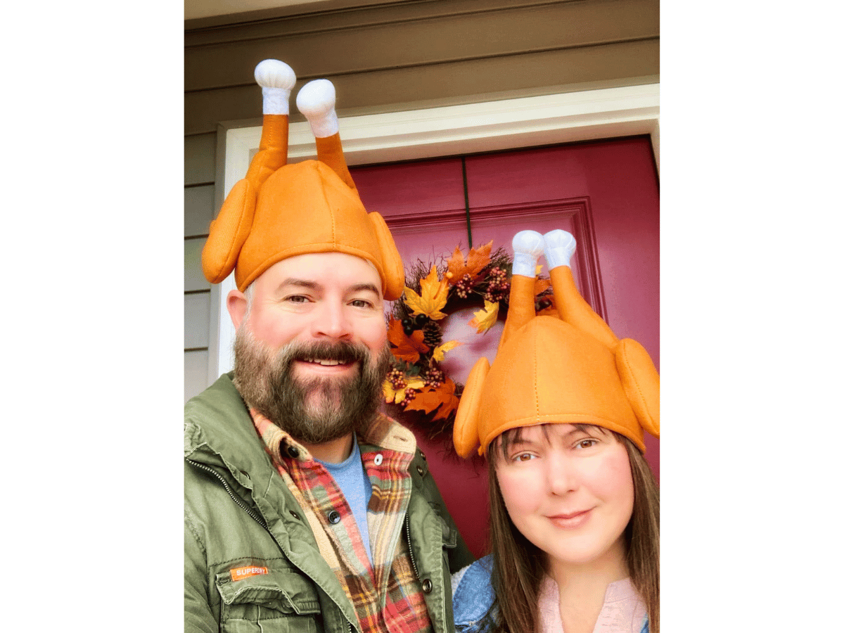 Couple smiling in orange turkey hats with white sock-like tips, posing at a front door with an autumn wreath behind them