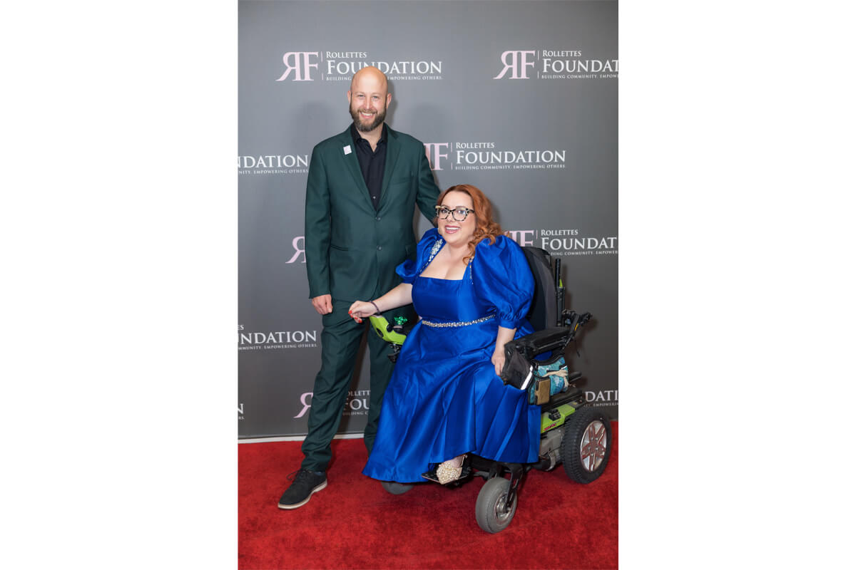 Man in a green suit stands beside a smiling woman in a royal blue gown seated in a motorized wheelchair on a red carpet at a charity event.