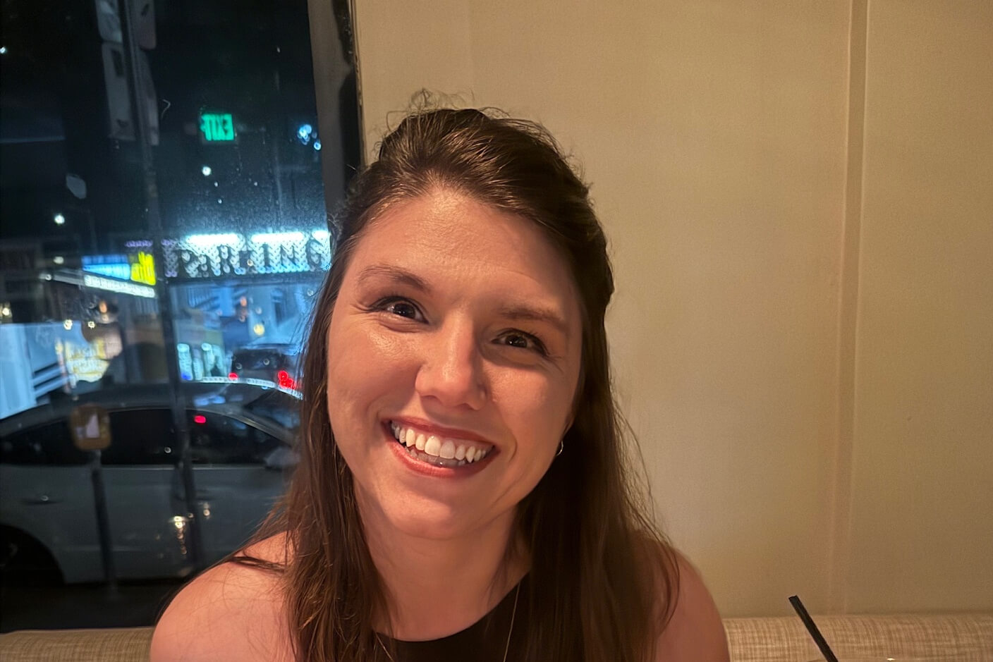 Close-up of a smiling woman with long brown hair sitting indoors by a window at night, city lights in the background outside.