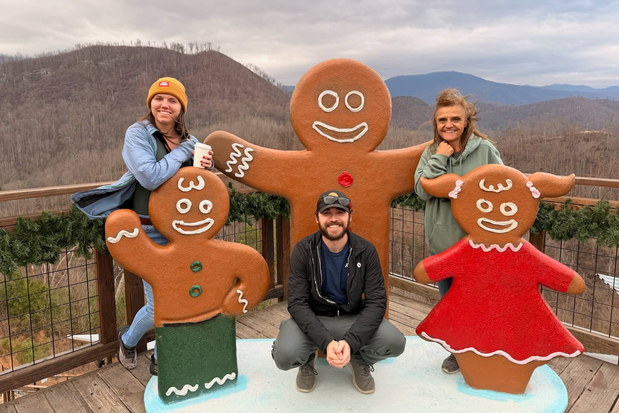 Three people posing with oversized gingerbread cookie sculptures at a mountain overlook, with bare trees and distant hills in the background.