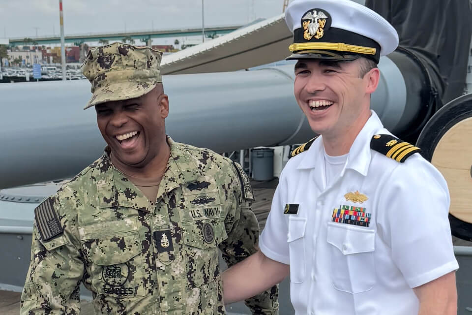 Two military officers laugh together on a ship deck, one in camouflage and one in a white Navy uniform with ribbons.