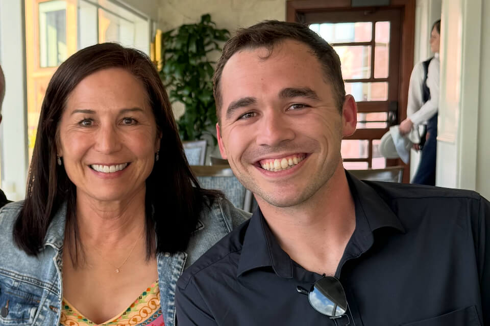 Two smiling people posing for a photo indoors at a bright cafe or restaurant; woman on the left wears a denim jacket and floral top, man on the right wears a black shirt with sunglasses hanging from his collar.