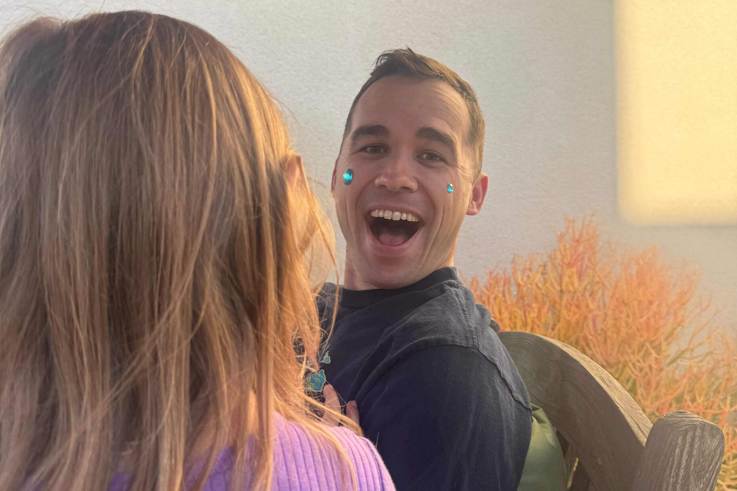 Smiling man taking a close-up selfie with turquoise face gems on his cheeks; person with long brown hair is in the foreground, outdoors with tall grasses in the background.