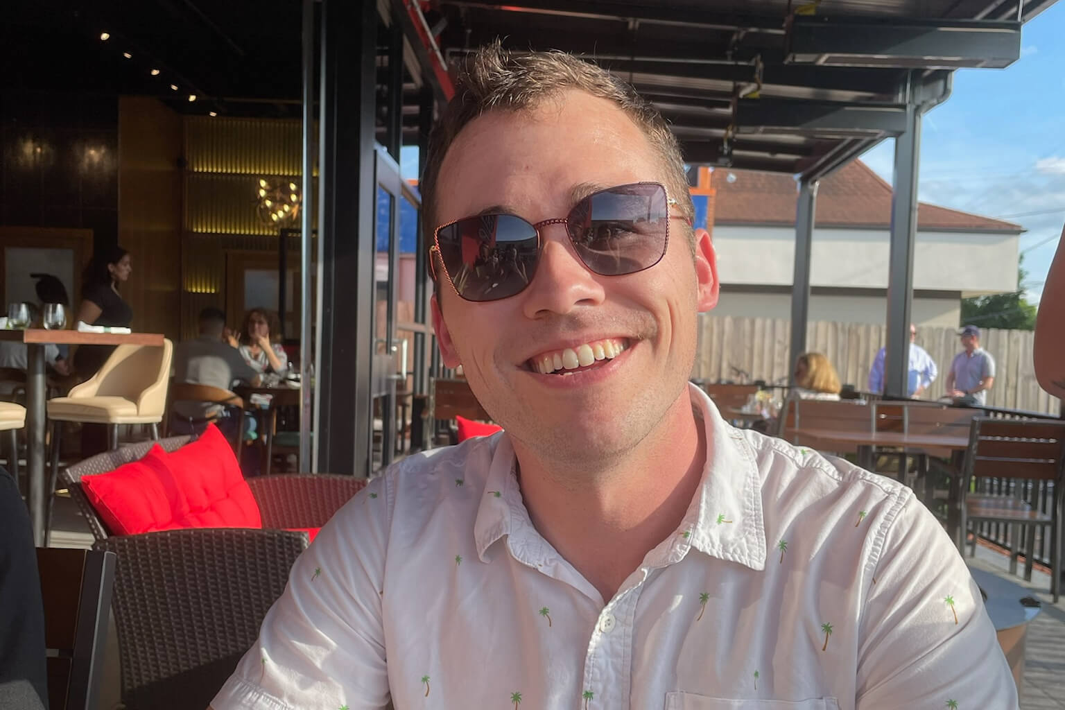 Man in sunglasses smiling at an outdoor restaurant table on a sunny day, wearing a white palm‑print shirt.