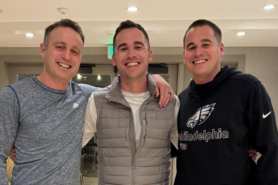 Three men stand side by side indoors, smiling for the camera, arms around each other.
