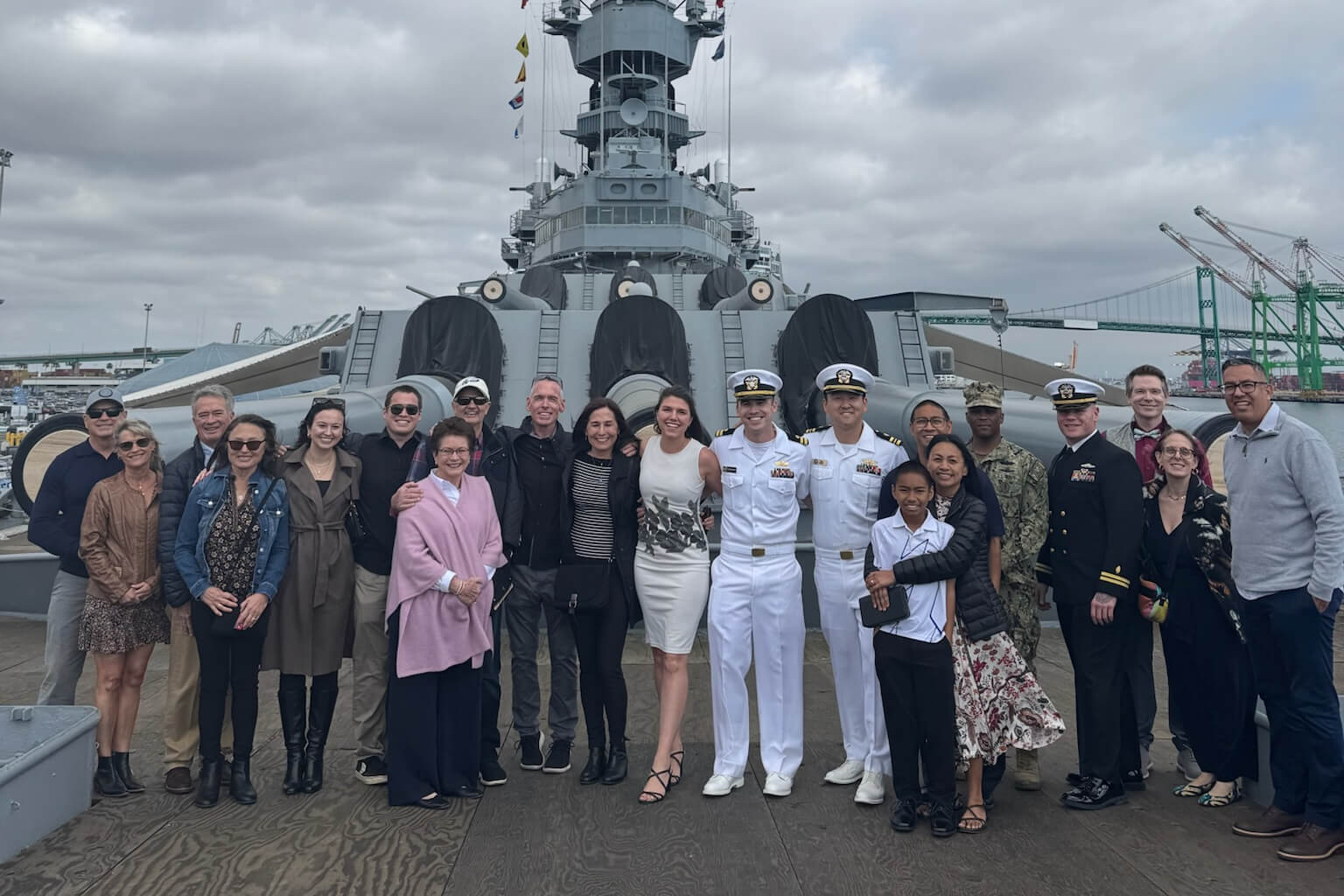Group photo of civilians and sailors on a ship's deck with a large battleship in the background.