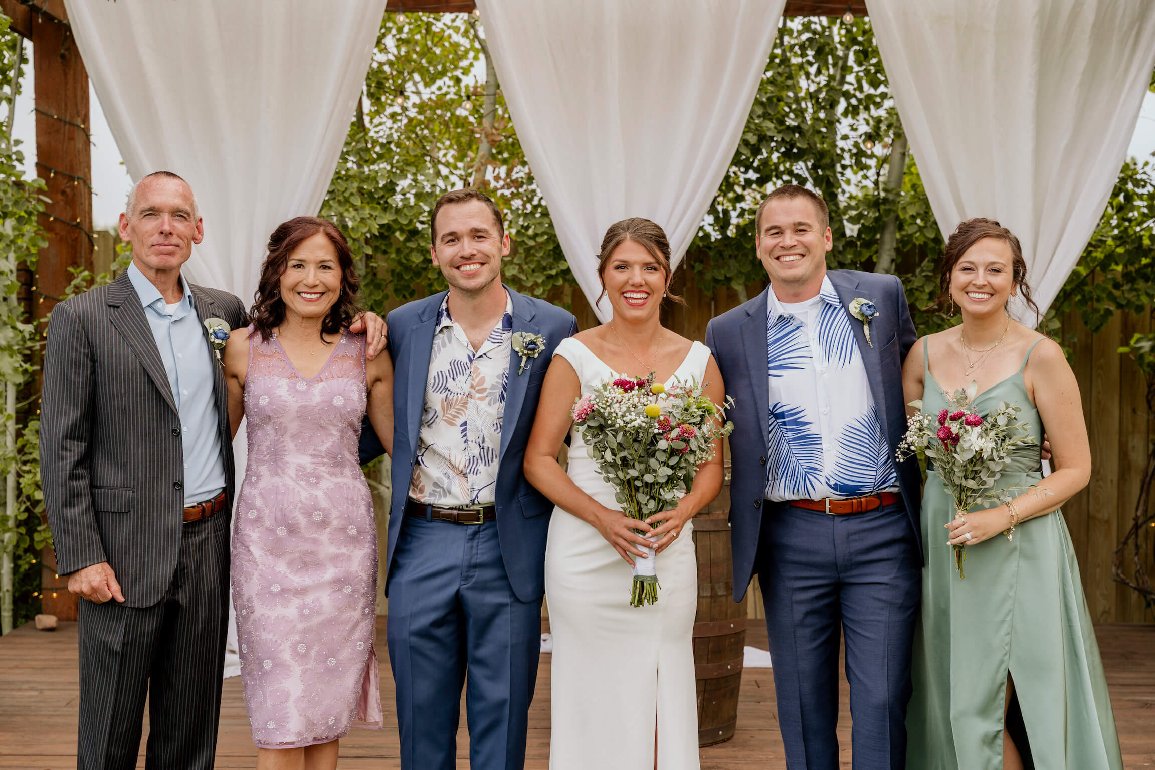 Wedding party posing outdoors under white drapery; six adults in formal attire, holding bouquets and smiling for a group photo.